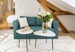 A teal sofa with white cushions, two white round tables with black legs, and a vase with white flowers.