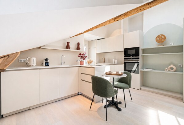 Una cocina moderna con armarios blancos, una zona de comedor con dos sillas verdes y un horno empotrado.