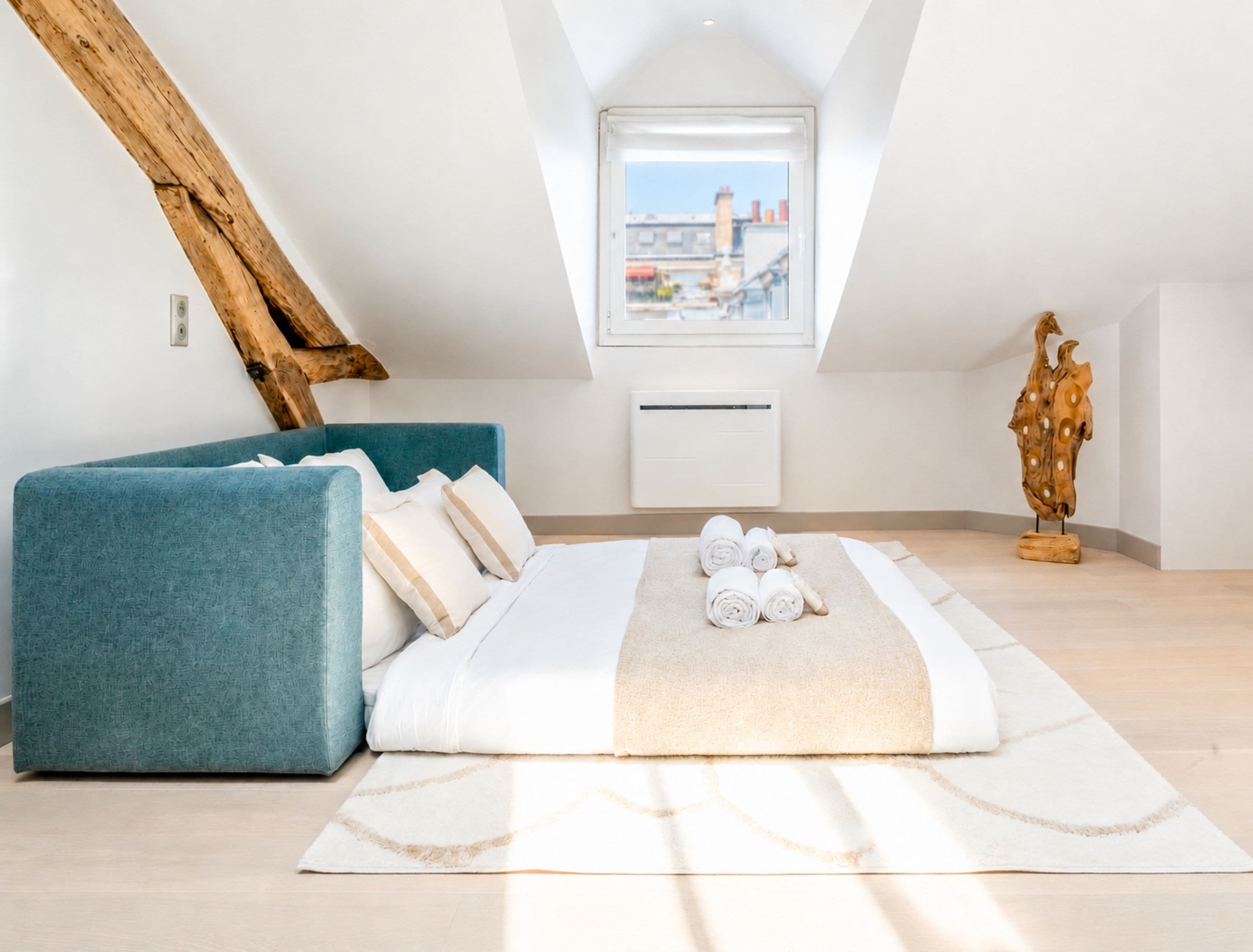 Un dormitorio con techo inclinado, una cama con toallas enrolladas, un sofá verde azulado y una escultura de madera.