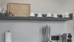 Een keuken met een houten kastje, een plank met witte bekers en glazen, een keukenrolhouder en een koffieapparaat.