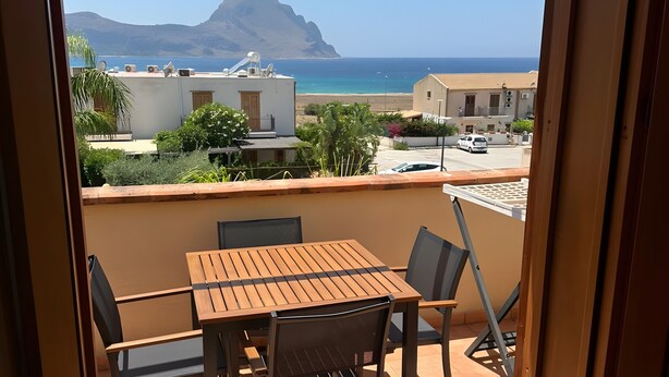 A balcony with a wooden table and chairs, overlooking a coastal view with buildings and a mountain.