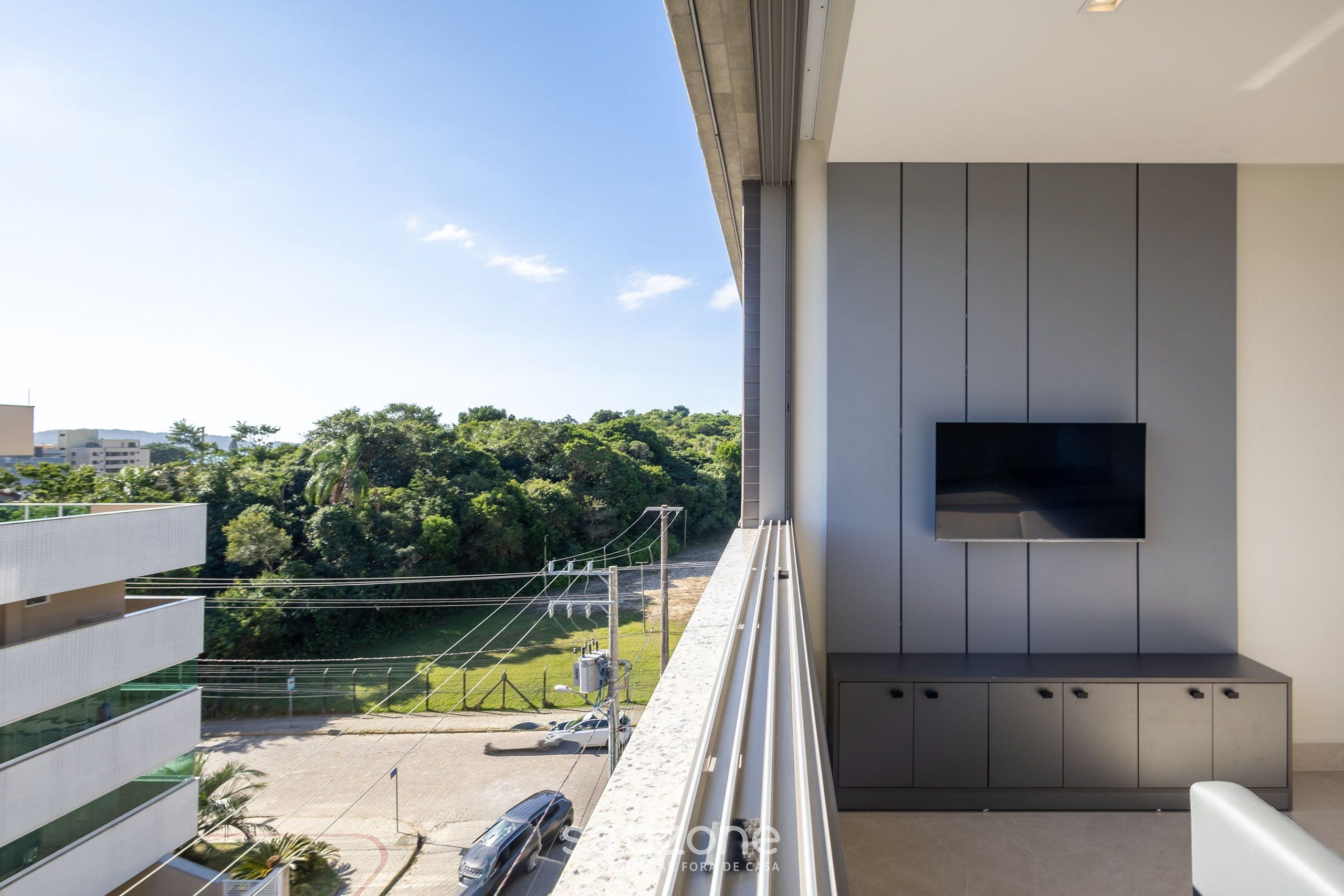 A balcony with a TV, a view of a parking lot, and greenery.