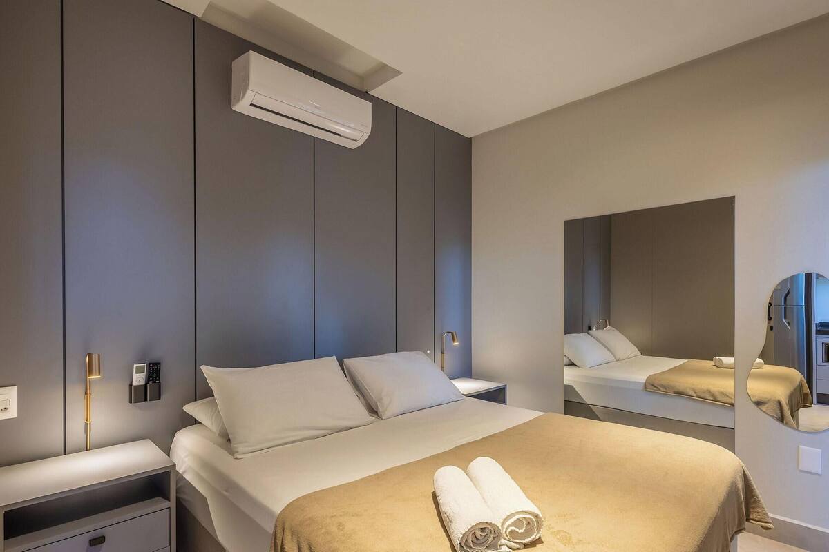 Une chambre d’hôtel moderne équipée d’un lit, de tables de chevet, d’un climatiseur, d’un miroir fixé au mur et d’une serviette posée sur le lit.