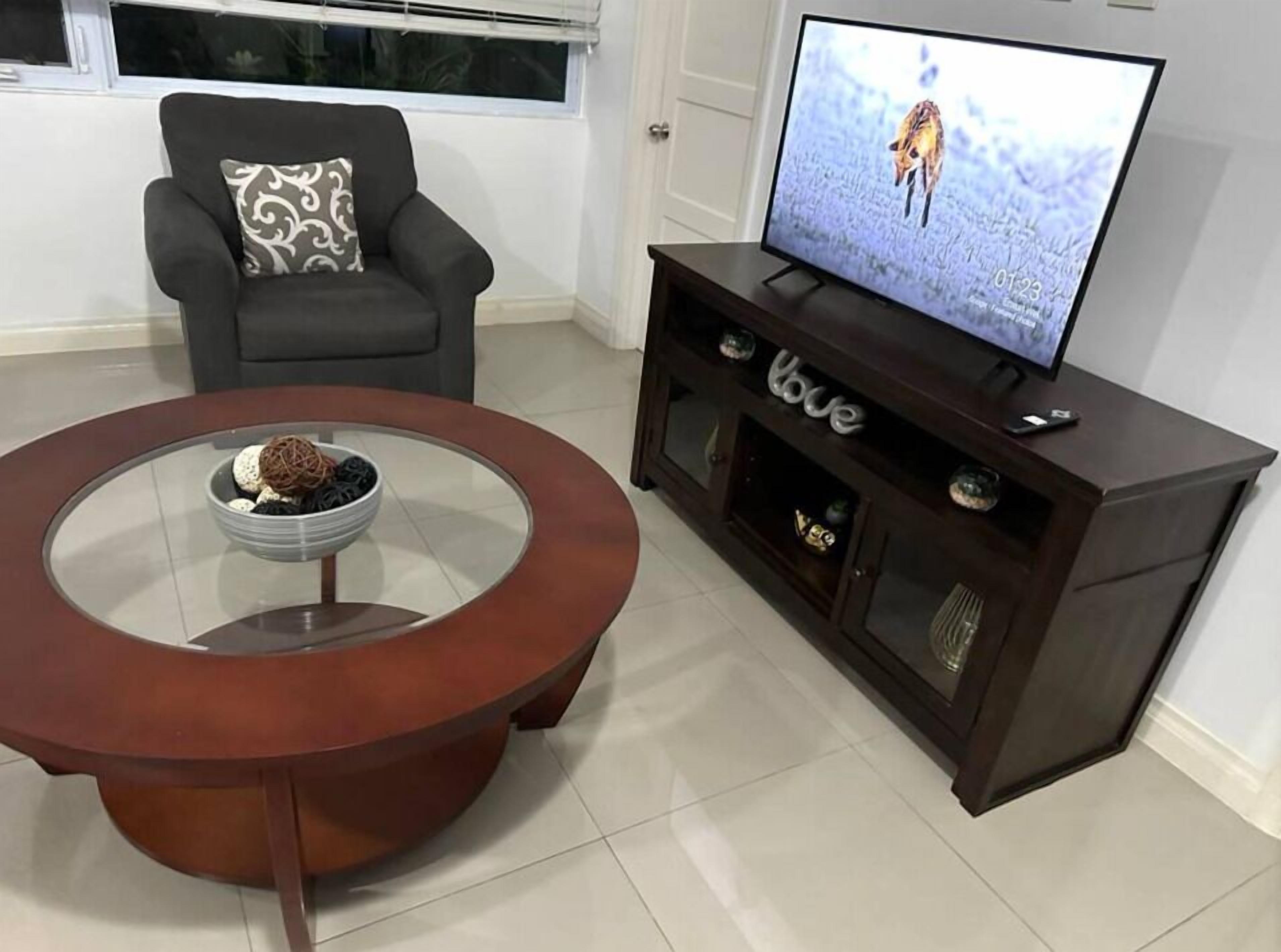 A living room with a round glass coffee table, a dark wooden TV stand with a flat-screen TV, a grey armchair with a patterned cushion, and a window with blinds.