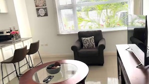 Ein modernes Wohnzimmer mit einem Couchtisch aus Glas, zwei Sesseln, einem Sofa, einem Fernseher und einer Küchenzeile mit Esstisch und Stühlen.