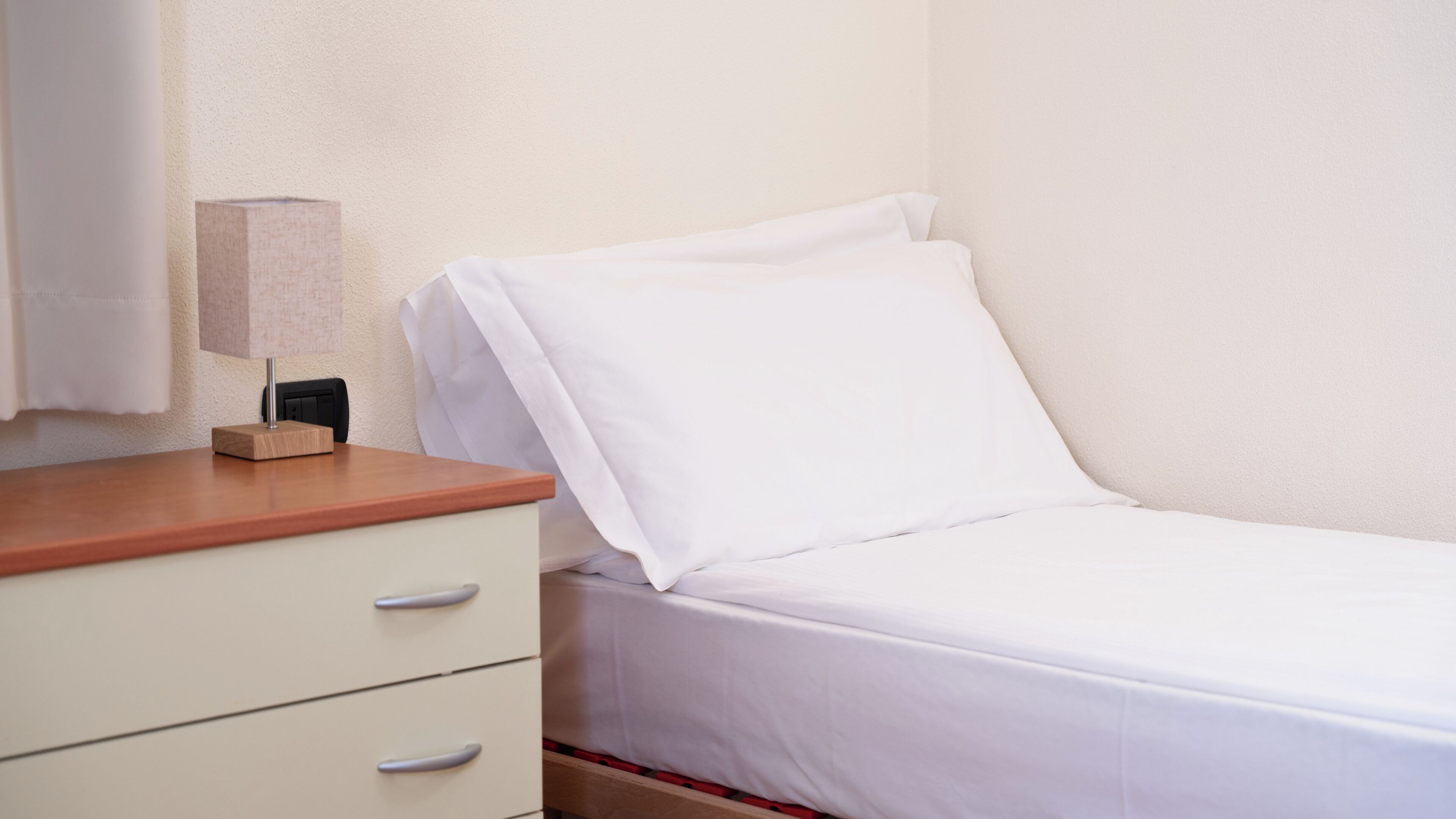 Una cama individual con ropa blanca de cama, una mesita de noche de madera, una lámpara y una pared blanca.