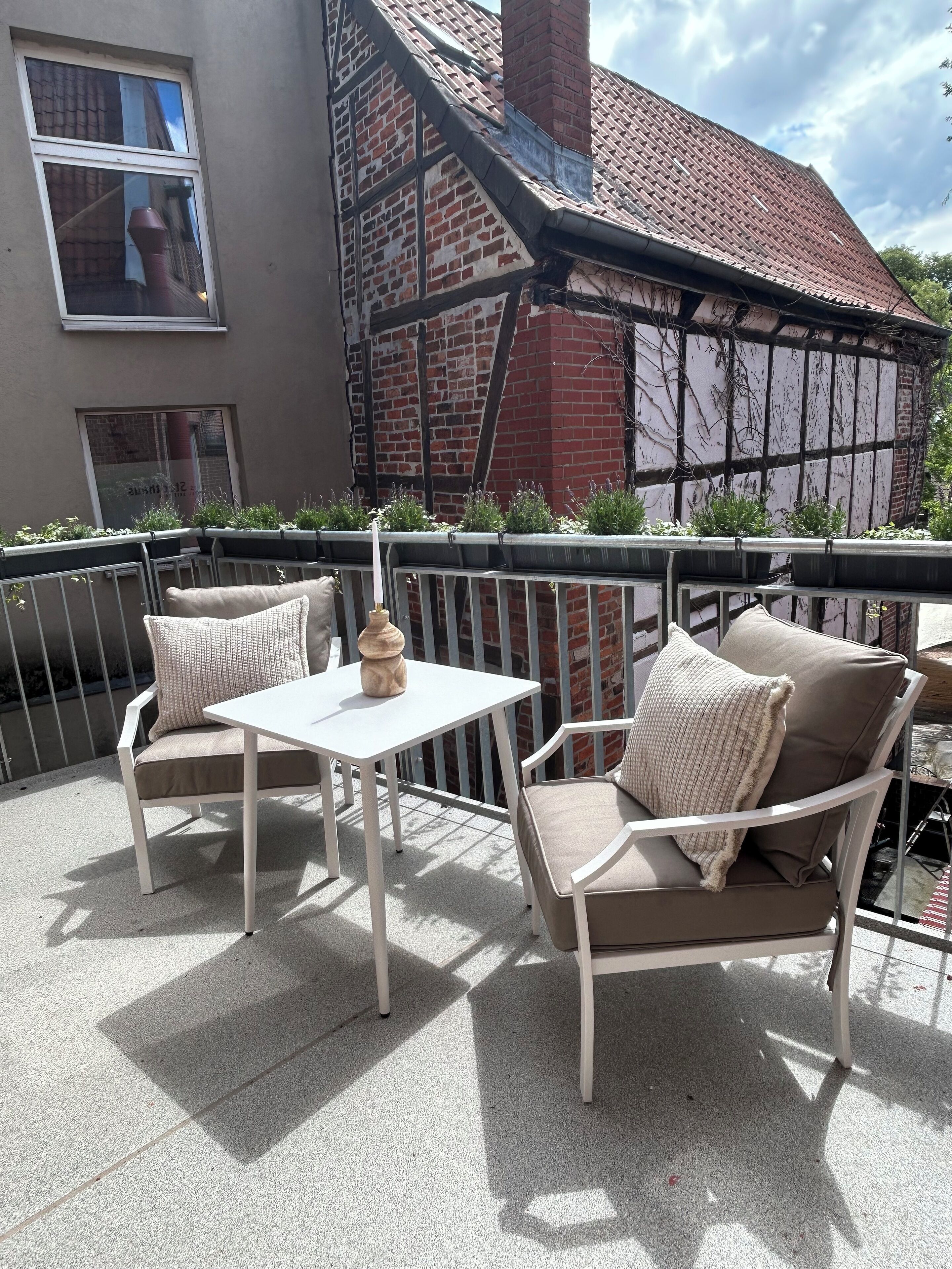 A balcony with a white table and two chairs, a brick wall, and a window.