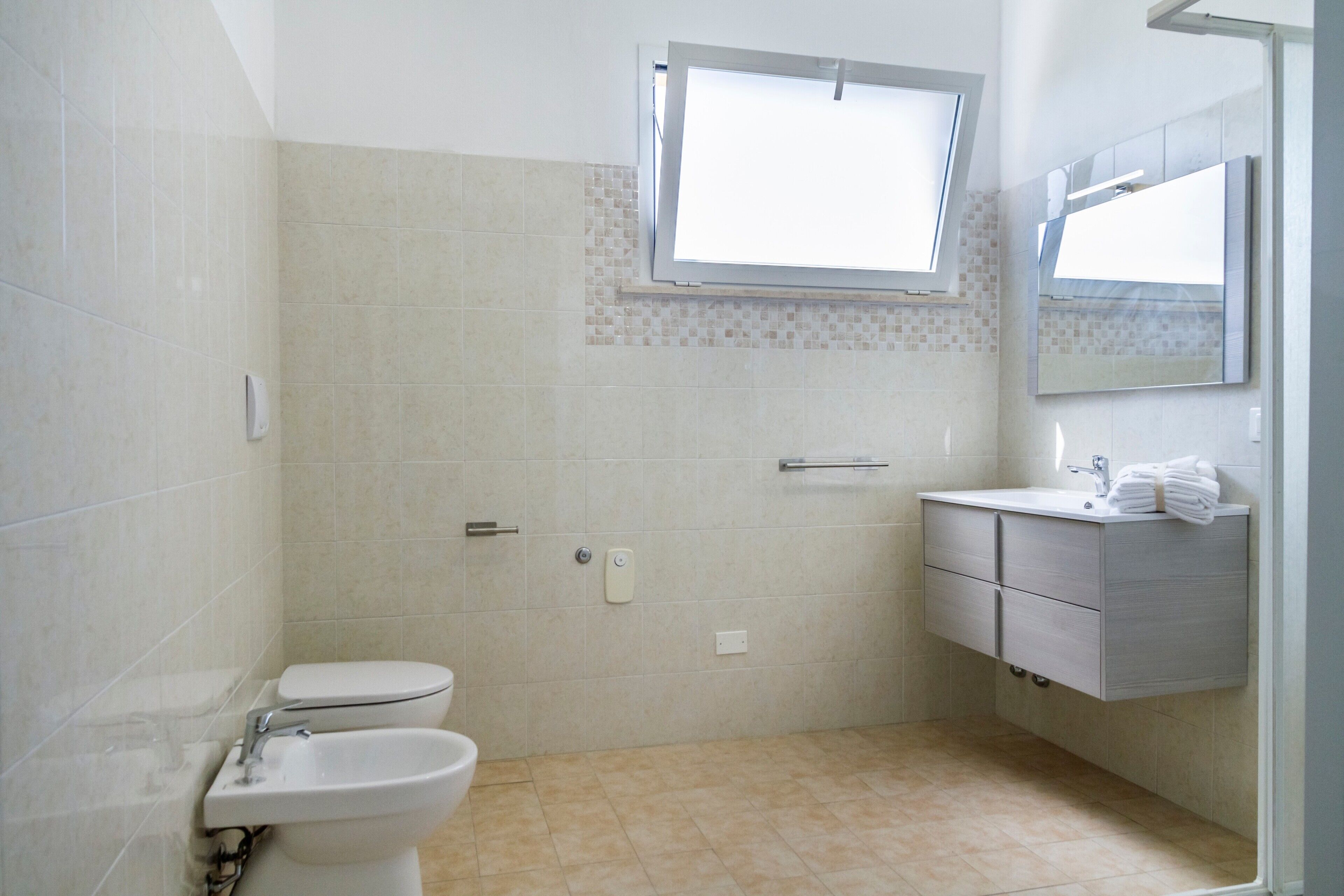 Une salle de bain équipée de toilettes, d’un lavabo et d’un meuble-lavabo, avec des murs carrelés et une fenêtre.
