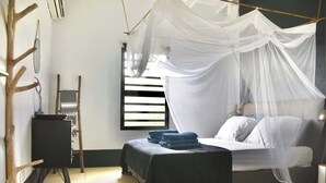 A room with a bed under a canopy, a small table with a chair, and a window with blinds.