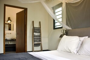 A bedroom with a bed, a nightstand, a bedside lamp, a window with curtains, and a bathroom visible through an open door.