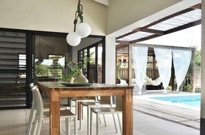 Un área de comedor al aire libre con una mesa y sillas de madera, una lámpara colgante y vistas a una piscina.