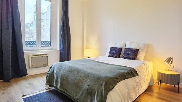 A bedroom with a bed, a window with curtains, a bedside lamp, and a radiator.