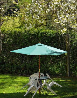 A table with a green umbrella, surrounded by white chairs, set on a grassy lawn with a hedge in the background. - Apart Hotel Rucaleufú (San Martín de los Andes)
