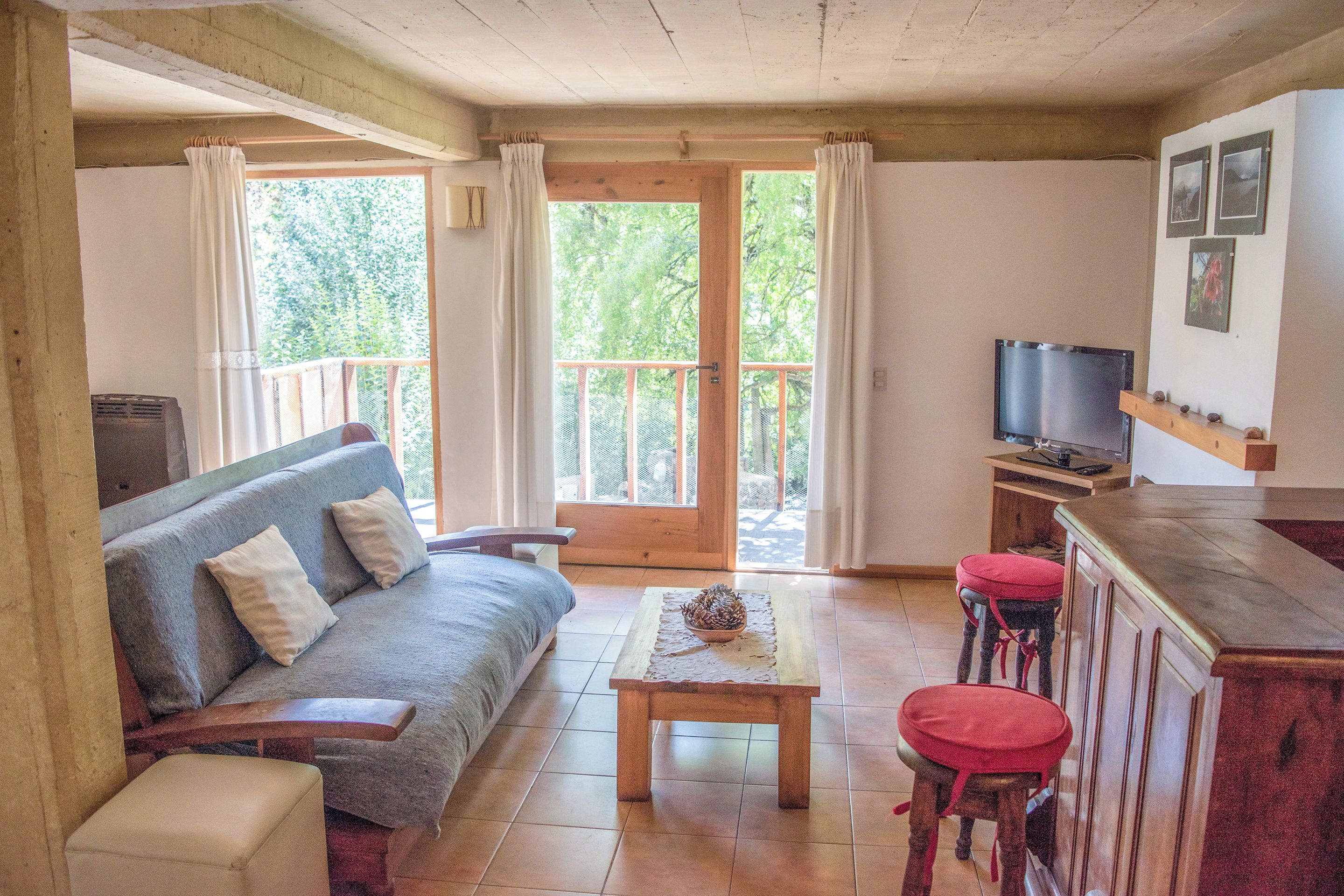 A cozy living room with a sofa, a coffee table, and a TV. There is a balcony with a wooden railing and a door leading outside.