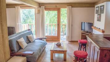A cozy living room with a sofa, a coffee table, and a TV. There is a balcony with a wooden railing and a door leading outside.
