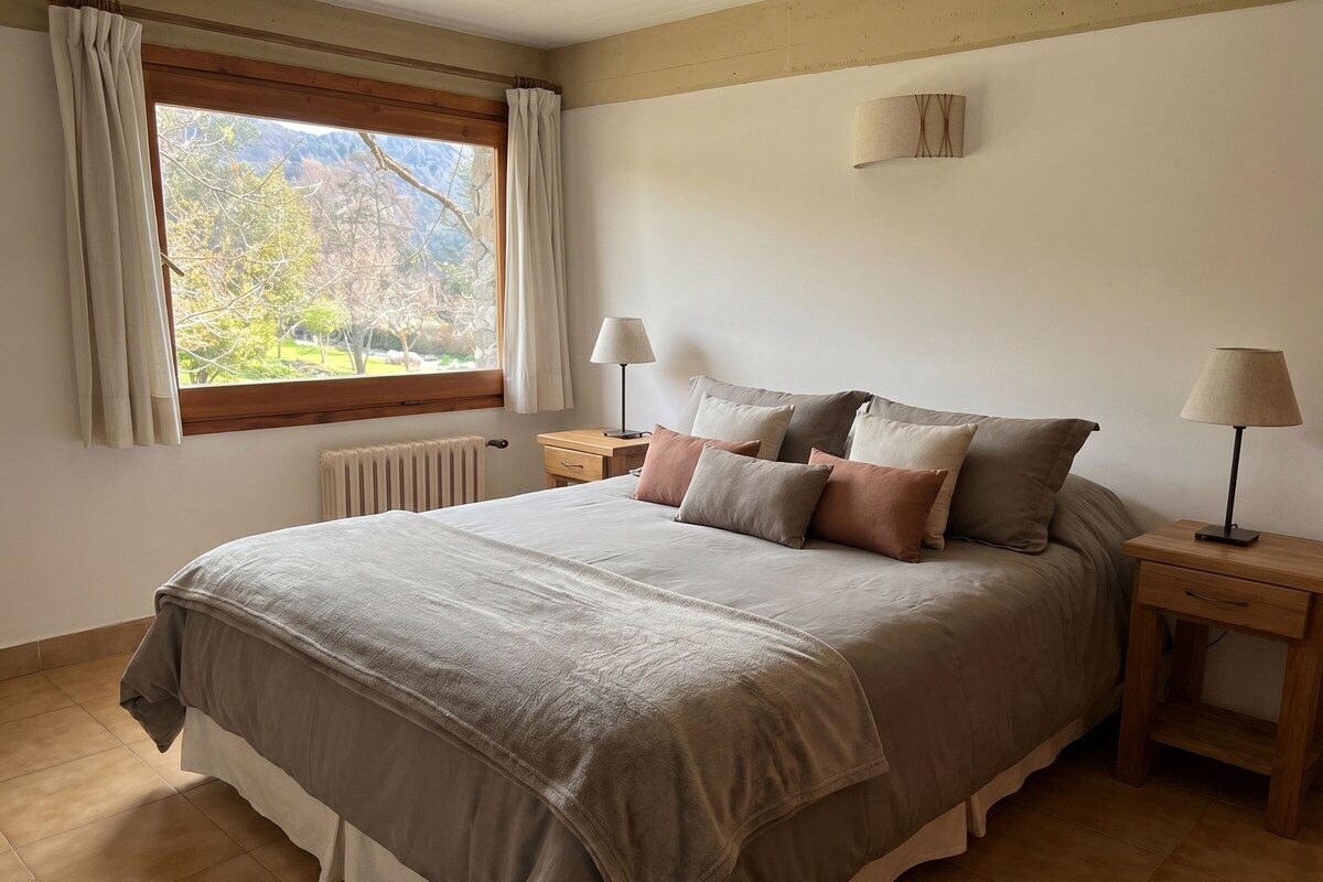 A bedroom with a bed, two bedside tables, a window with curtains, a radiator, and a lamp.