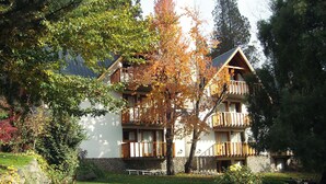 A multi-story building with balconies, surrounded by a well-maintained garden with a stone wall and a variety of trees. - Apart Hotel Rucaleufú (San Martín de los Andes)