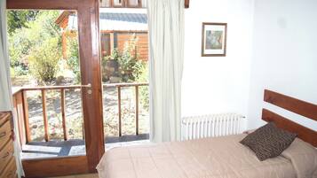 Une chambre à coucher comprenant un lit, une tête de lit en bois, une fenêtre donnant sur un jardin, un radiateur et une porte menant à un espace extérieur.
