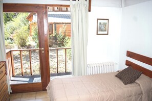 A bedroom with a bed, a wooden headboard, a window with a view of a garden, a radiator, and a door leading to an outdoor area. - Apart Hotel Rucaleufú (San Martín de los Andes)