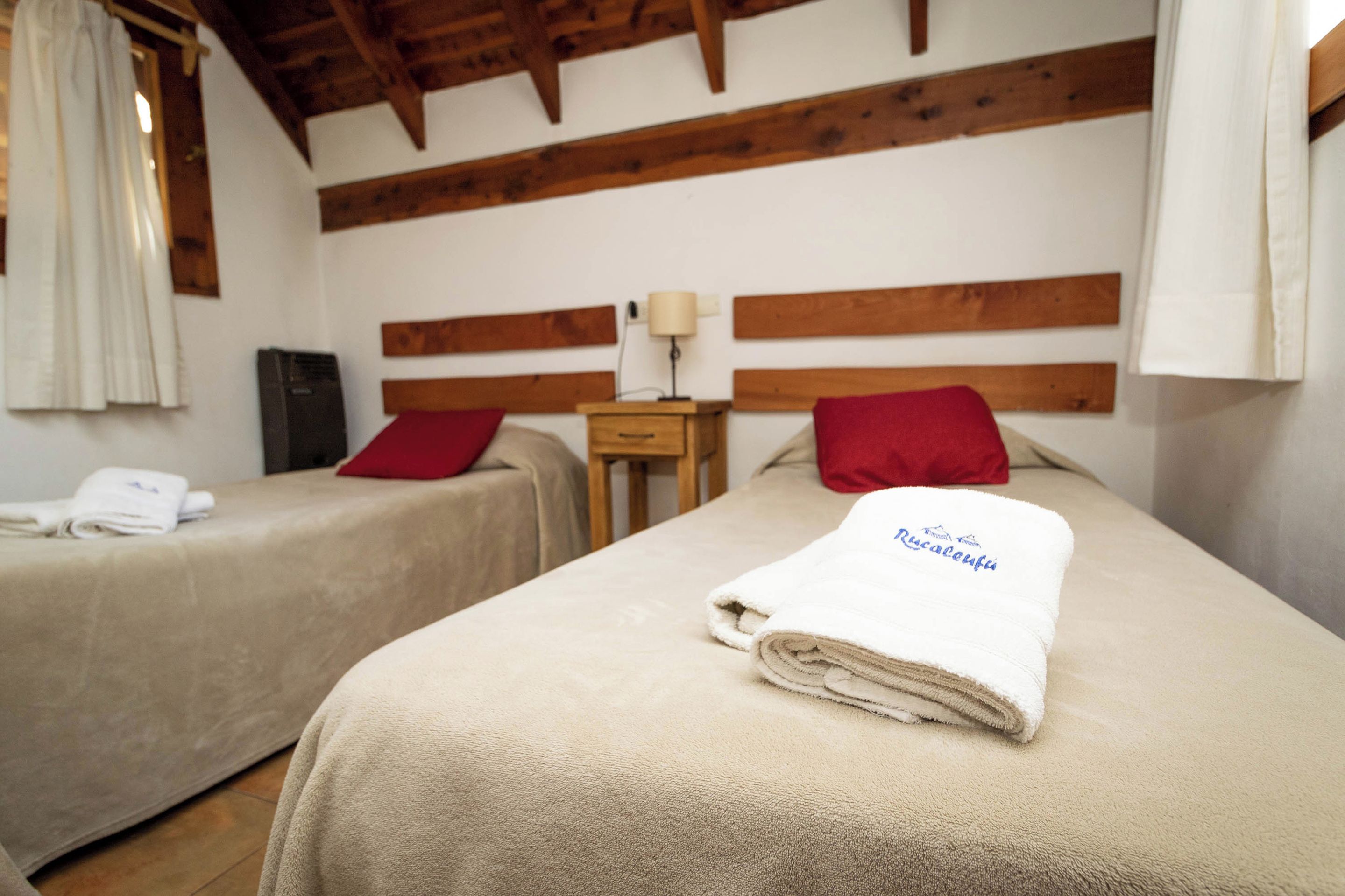Una habitación con dos camas, cada una con una almohada roja y una toalla blanca. Entre las camas hay una mesita de noche con una lámpara.