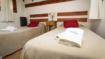 Una habitación con dos camas, cada una con una almohada roja y una toalla blanca. Entre las camas hay una mesita de noche con una lámpara.