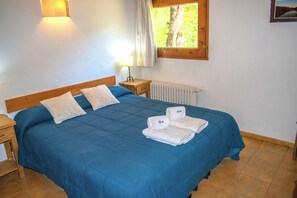 A neatly made bed with blue bedding, a bedside table with a lamp, a window with curtains, and a radiator. - Apart Hotel Rucaleufú (San Martín de los Andes)