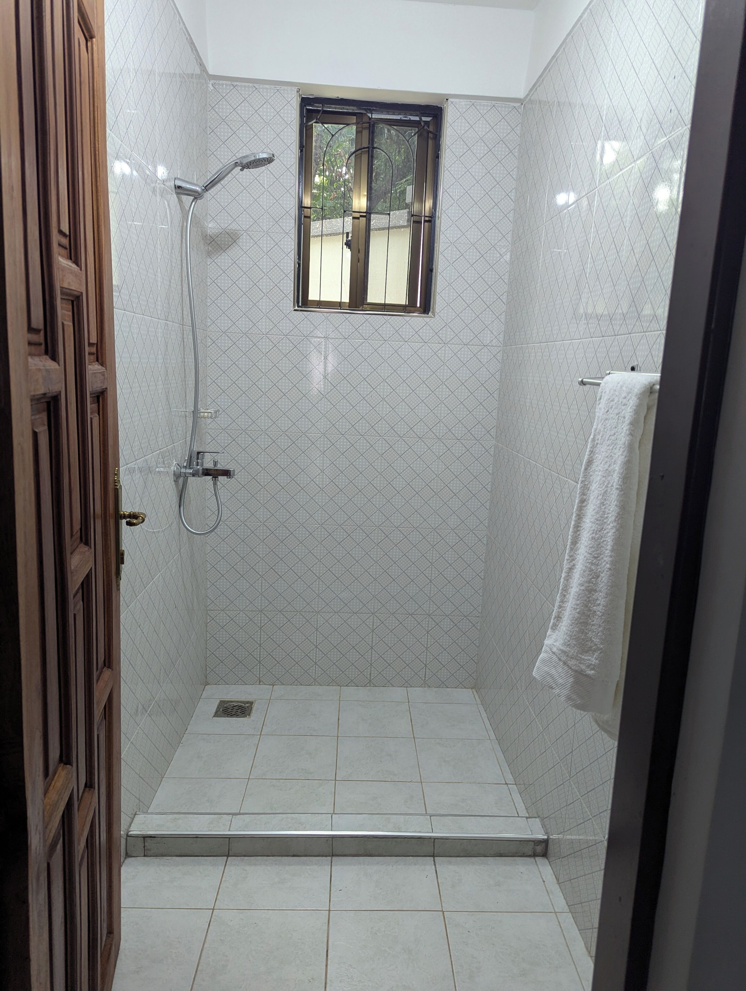 A small, tiled shower room with a window, a towel rack, and a wooden door.