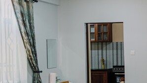 A dining area with a wooden table and chairs, a kitchen in the background, and a window with curtains. - St. Benedict House (Moshi)