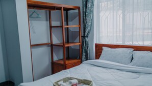 A neatly made bed with a basket of toiletries, a wooden wardrobe, and a window with curtains. - St. Benedict House (Moshi)