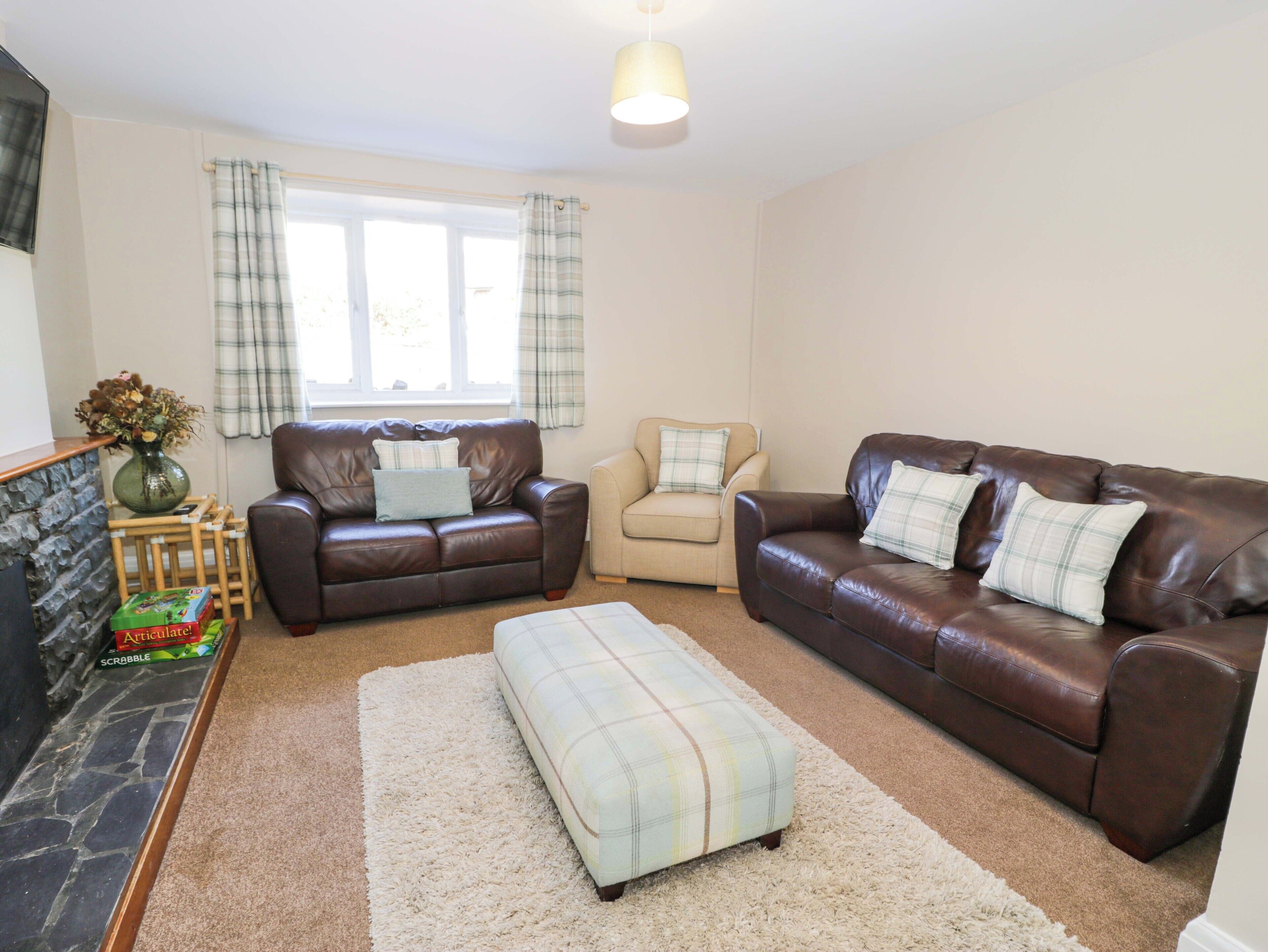 A living room with two leather sofas, a beige armchair, a plaid ottoman, a stone fireplace, and a window with curtains.