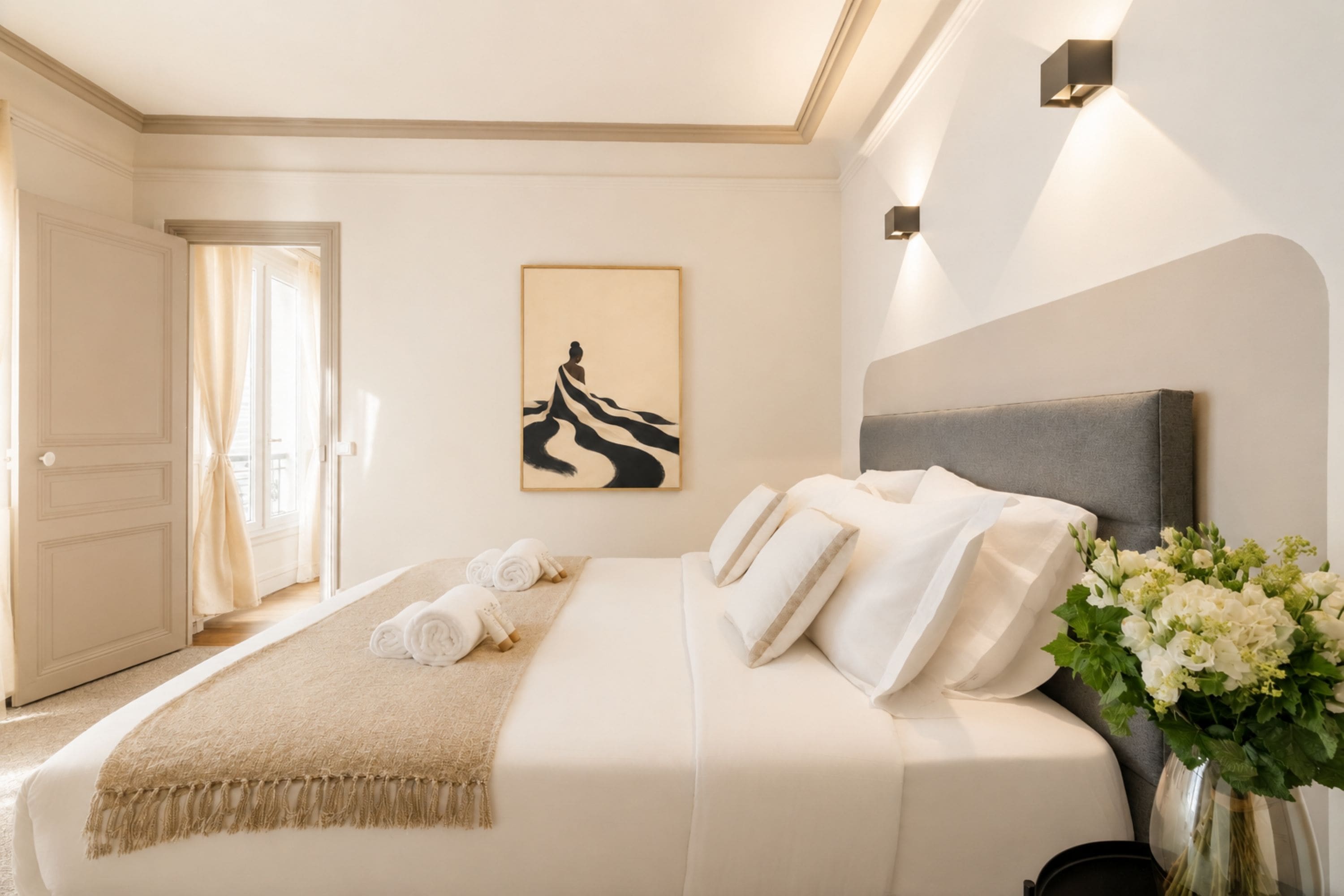 Hotellihuone, jossa on sänky, taulu, maljakko kukkineen ja ovi, joka johtaa toiseen huoneeseen.