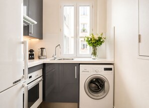 A compact kitchen with a built-in washing machine, a sink, and a window above it. - 1539 Belles Feuilles (Paris)