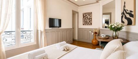 A hotel room with a large bed, a flat-screen TV, a decorative wall piece, a vase with flowers, and a view of a balcony.