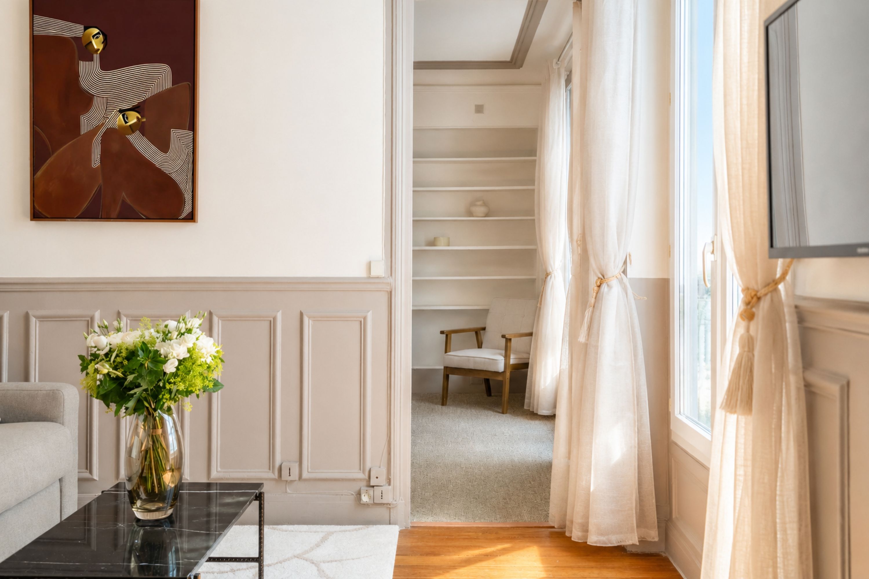 Una sala de estar moderna con un sofá, una mesa de centro con un florero, un cuadro en la pared y vistas a un dormitorio a través de un vano.