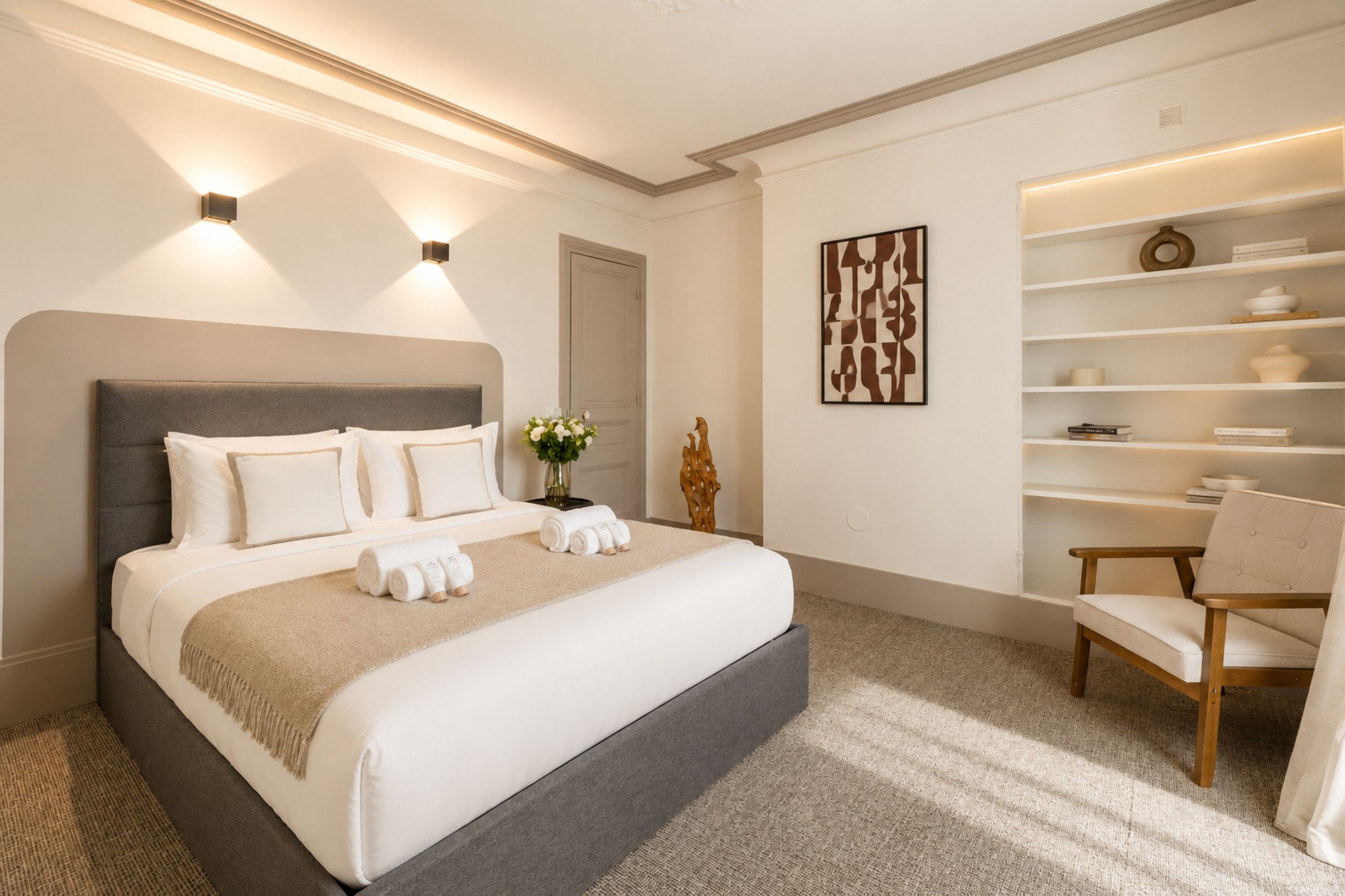 Un dormitorio moderno con una cama grande, lámparas de noche, un elemento decorativo en la pared y un estante empotrado con libros y objetos decorativos.