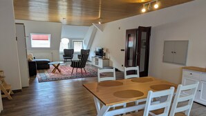 A spacious living area with a wooden ceiling, a dining table with white chairs, a sofa, a TV, and a cabinet. - A vacation with a view, a pleasant break in the heart of the city Dogs welcome (Bad Lauterberg)