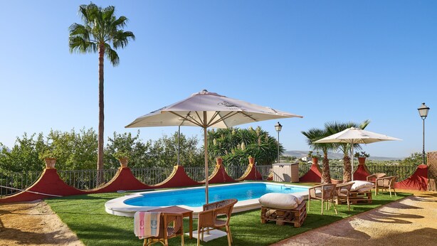 A swimming pool with lounge chairs and umbrellas, surrounded by palm trees and a red fence.