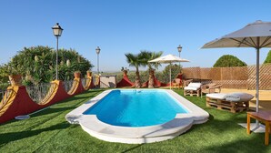 A rectangular swimming pool with a white deck, surrounded by artificial grass, palm trees, and outdoor furniture.