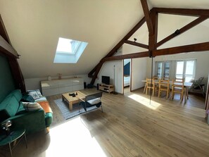 Un loft espacioso con techo alto, vigas de madera, tragaluces, zona de comedor con sillas de madera, un sofá y un televisor.
