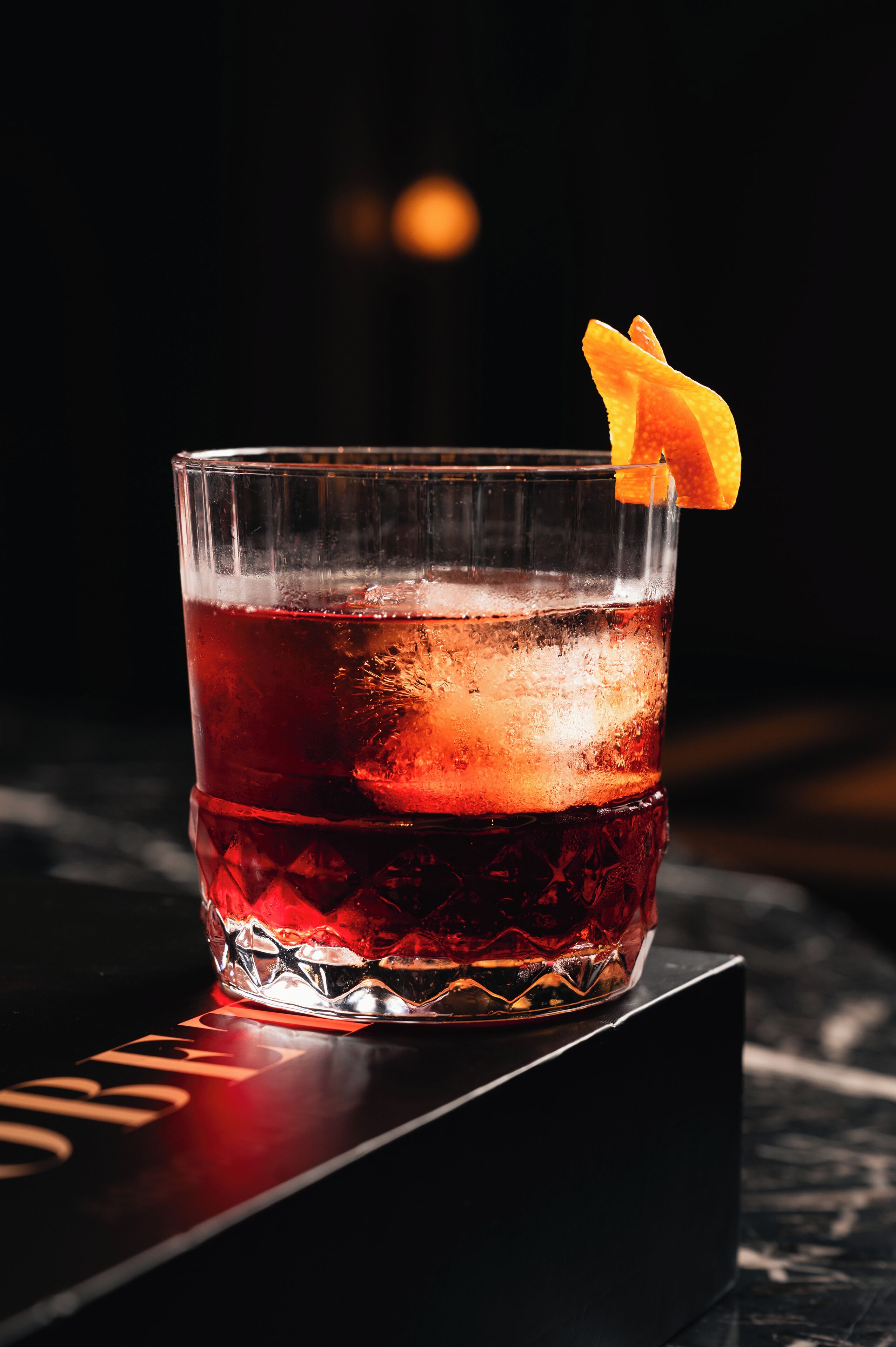 A glass of red beverage with ice cubes and an orange garnish on a black coaster.
