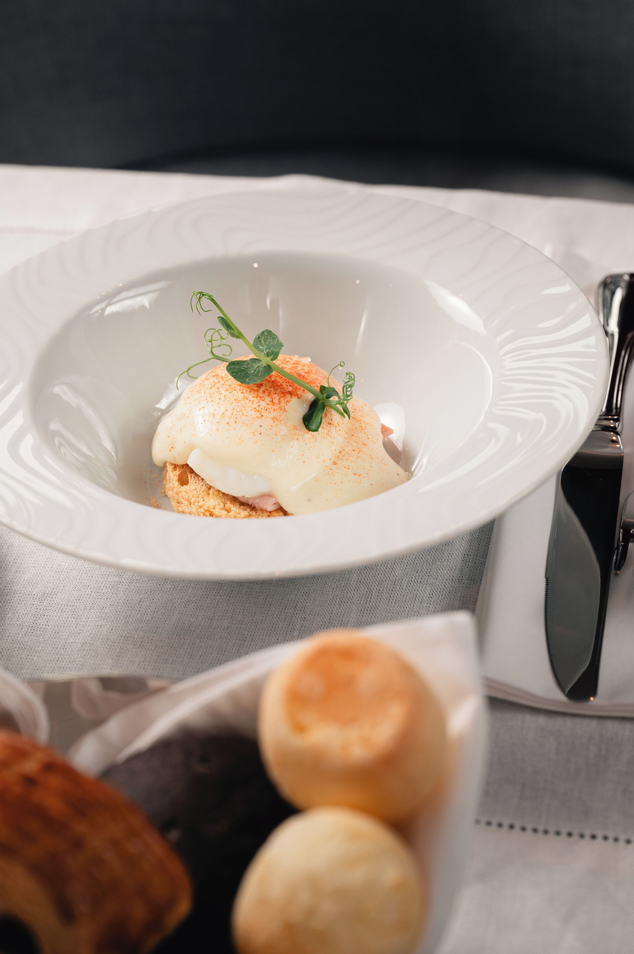 A white plate with a poached egg on toast, garnished with a sprig of parsley, and a small bowl of bread rolls in the foreground.