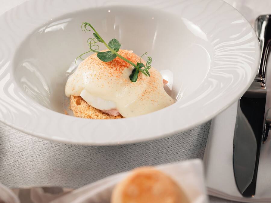 A white plate with a poached egg on toast, garnished with a sprig of parsley, and a small bowl of bread rolls in the foreground.