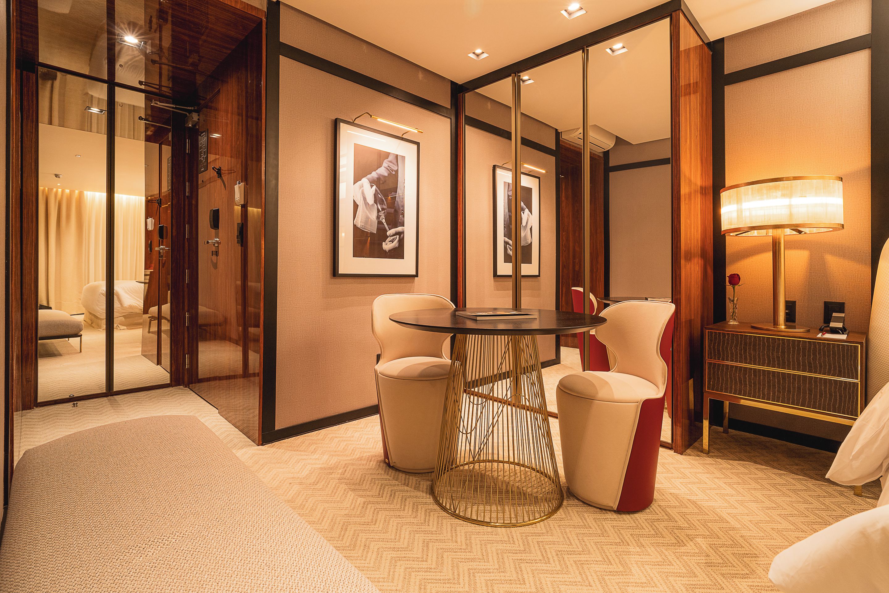 A hotel room with a glass partition, a round table, two chairs, a red accent chair, a lamp, and framed pictures on the wall.