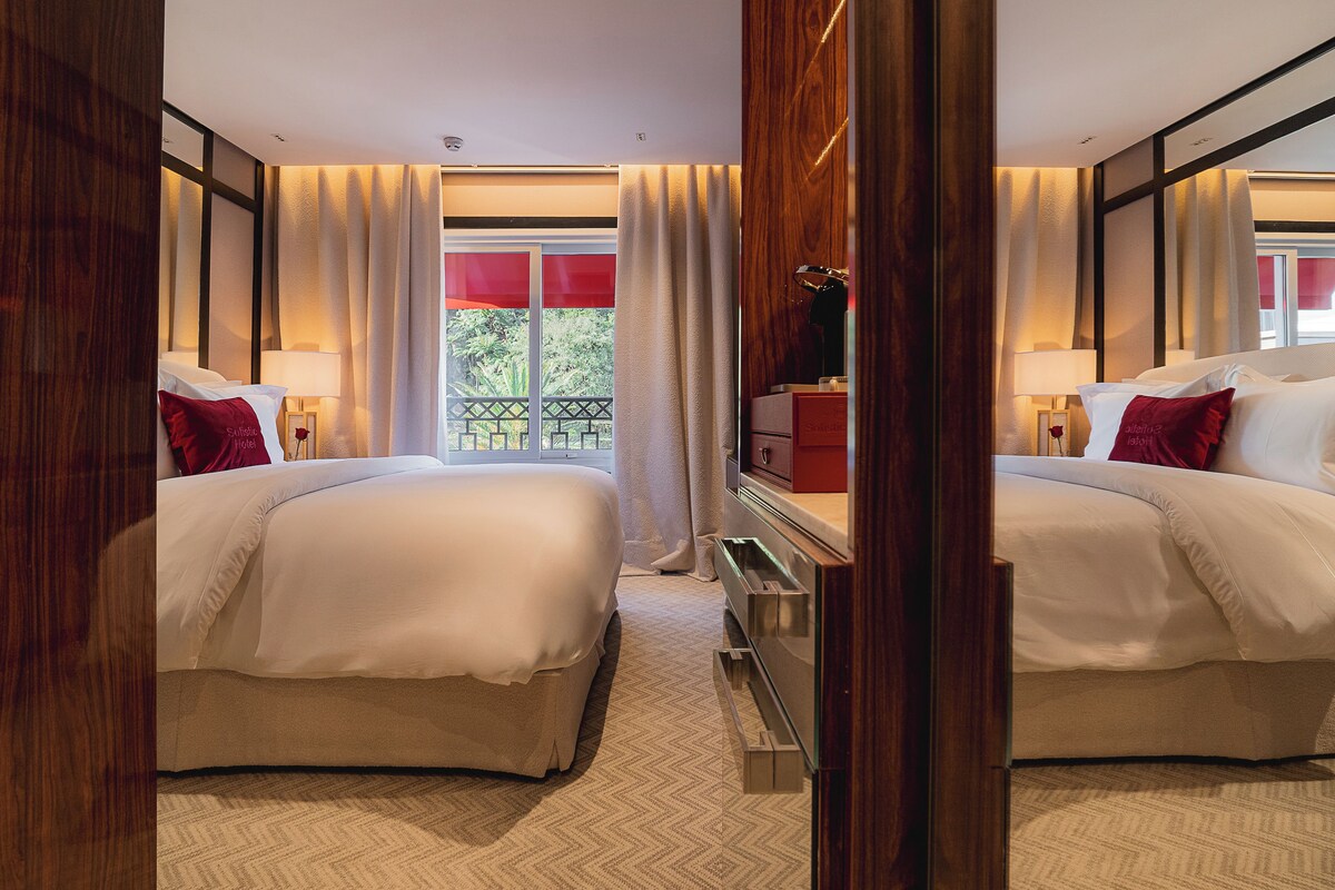 Quarto de hotel com duas camas, cômoda, espelho e janela com cortinas.
