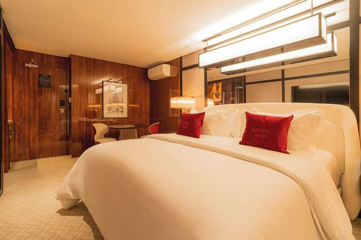 A hotel room with a large bed, a wooden wardrobe, a desk with a chair, and a mirror.