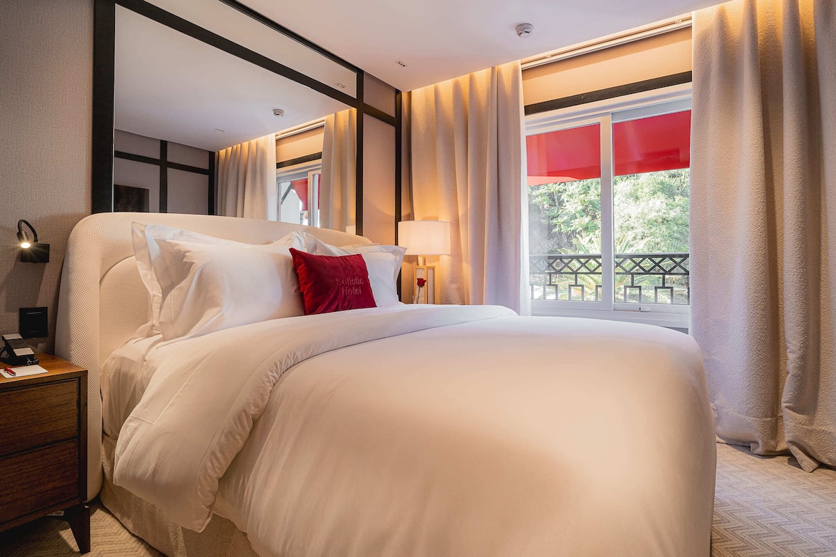 A hotel room with a large bed, a nightstand, a lamp, and a view of the outdoors.