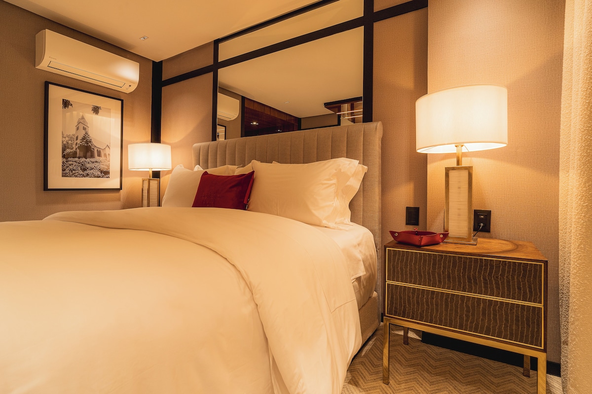 A hotel room with a large bed, a bedside table with a lamp, a framed picture on the wall, and an air conditioning unit.