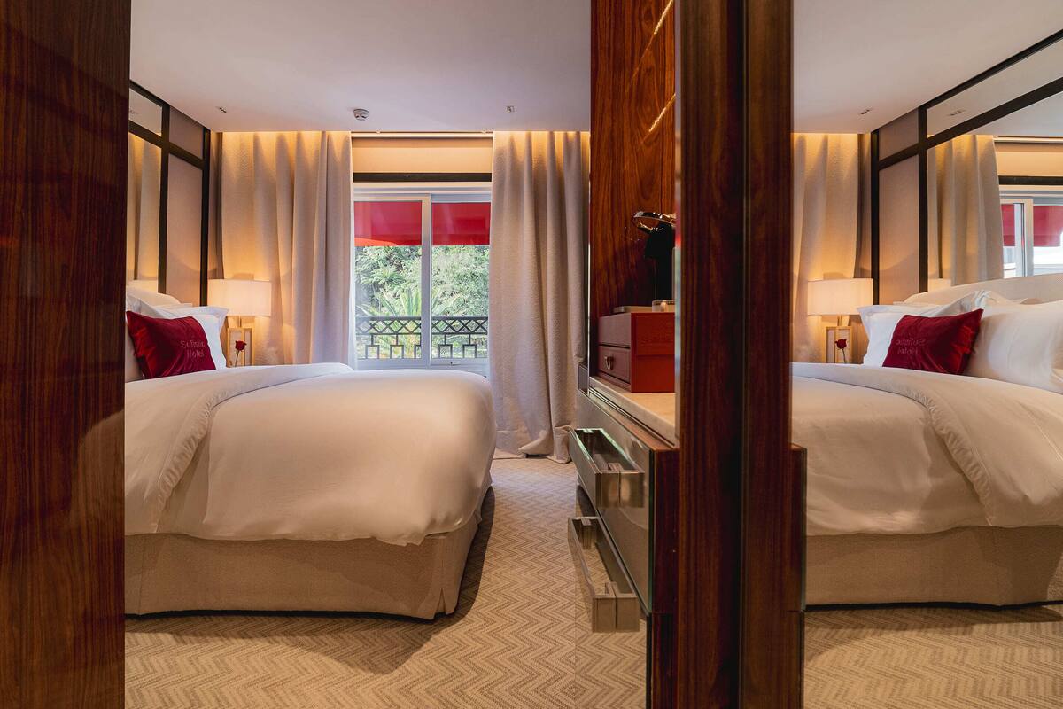 A hotel room with two beds, a wooden wardrobe, a small desk, and a window with curtains.