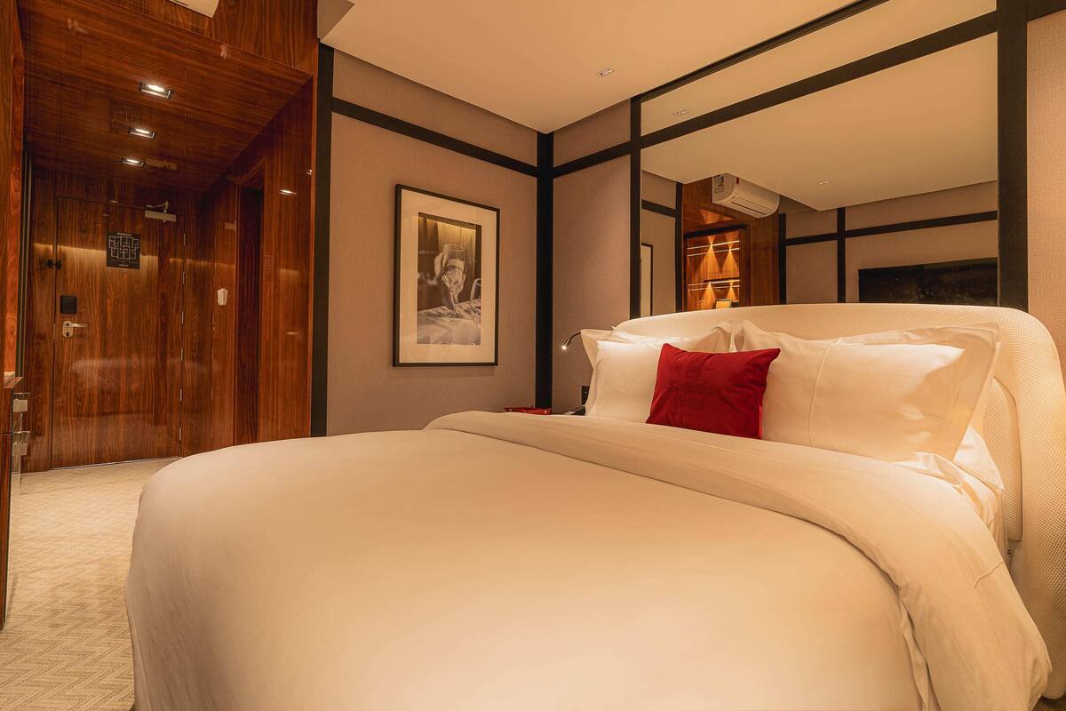 A hotel room with a large bed, a wooden wardrobe, a framed picture on the wall, and a mirror reflecting the room.