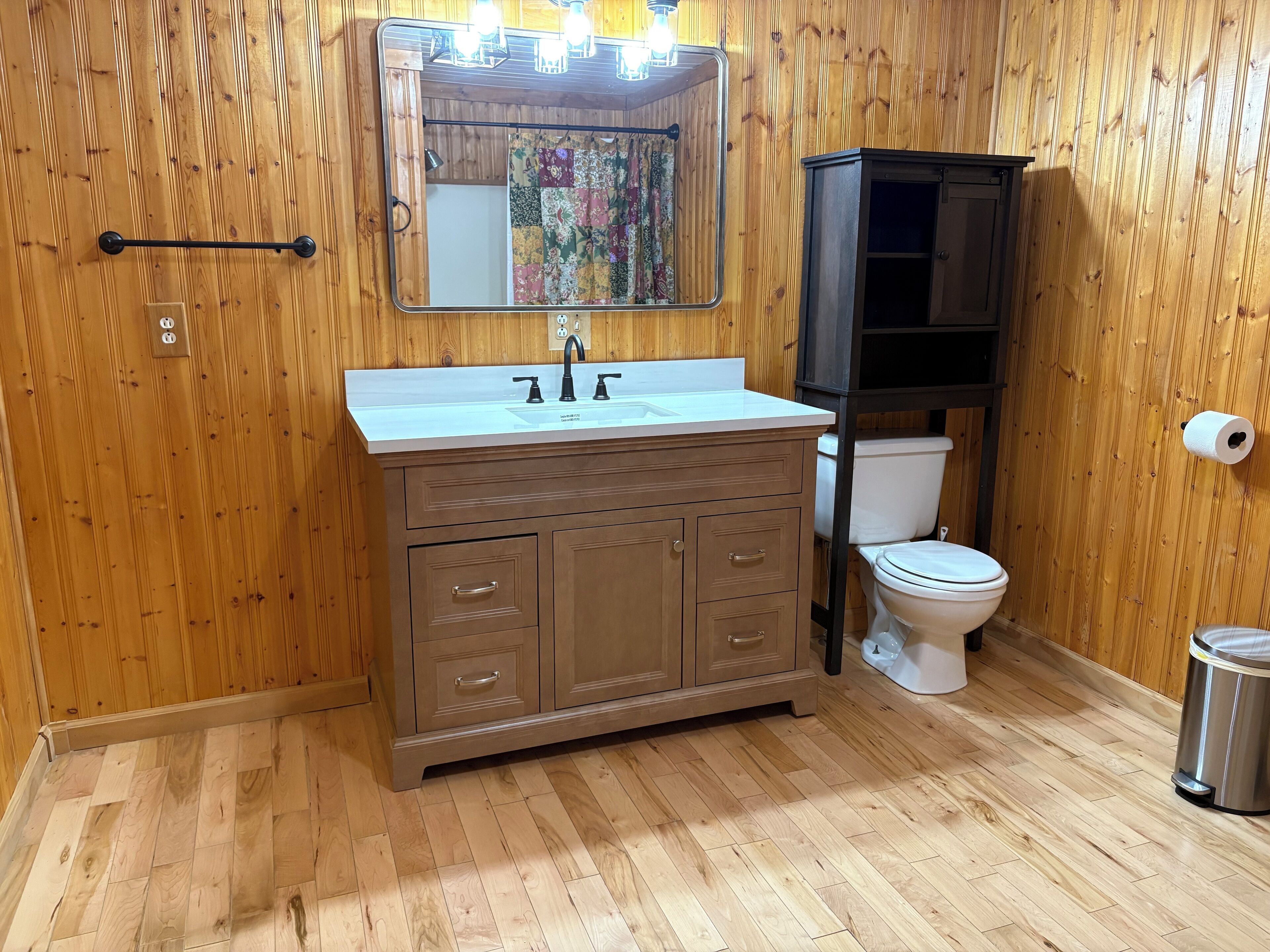 Une salle de bain aux murs lambrissés, avec un meuble-lavabo en bois, un évier, un miroir, des toilettes et une poubelle.